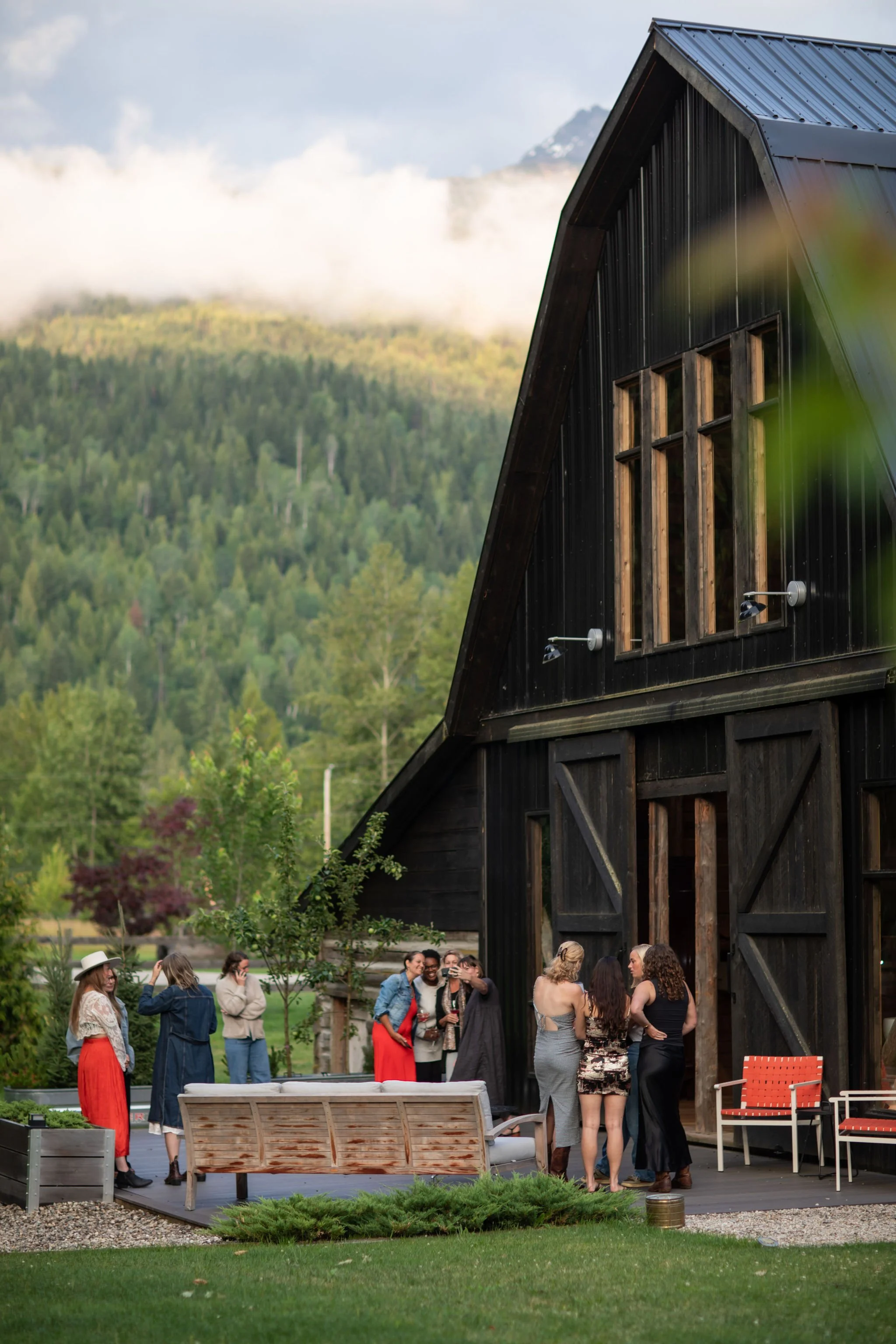 Group of people socializing outside near a black barn in a rural mountainous area with trees and clouds in the background.