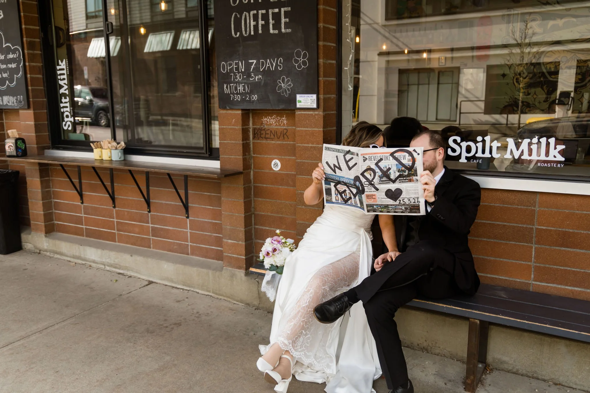 couple in wedding dress and tuxedo read a newspaper that says we eloped outside of a coffee shop