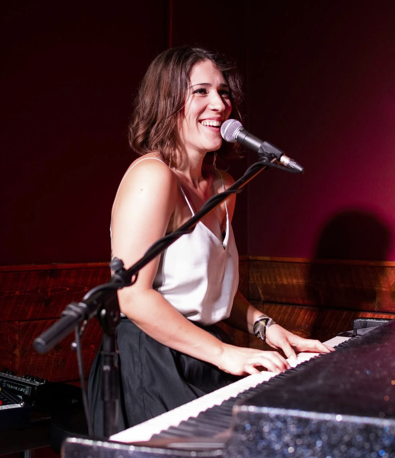A woman with shoulder-length brown hair smiling and singing into a microphone while playing a piano in a dimly lit room.