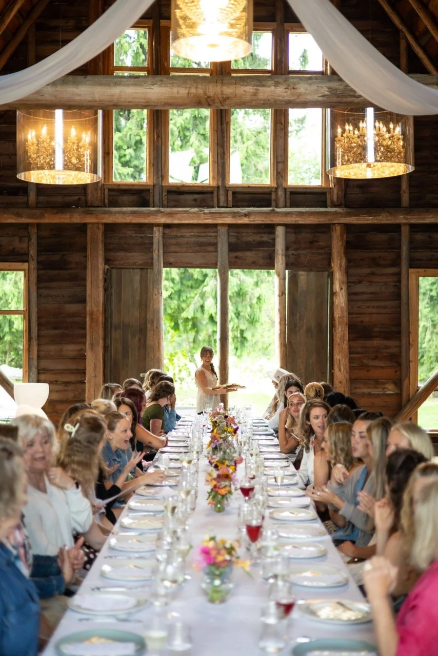 A large rustic wooden barn with a high ceiling, decorated for a wedding reception or special event, featuring a long banquet table with floral centerpieces, surrounded by seated guests, and a woman standing at the end of the table holding a platter.