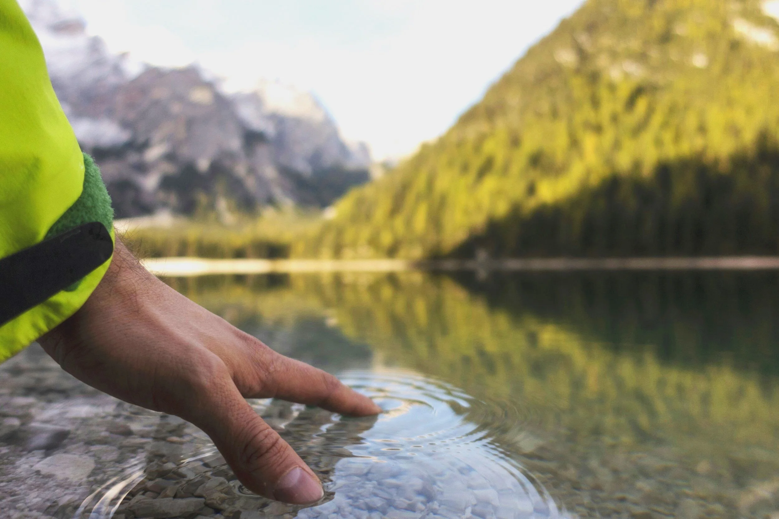 Close-up of a hand with fingers submerged in clear water near a rocky shoreline, with a scenic view of mountains and forest reflected in the lake in the background.