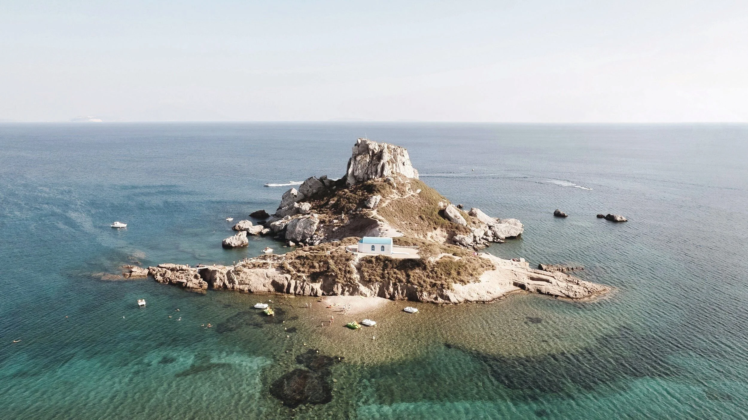 A small rocky island with a white building featuring a blue roof, surrounded by clear blue water with boats docked or floating nearby.