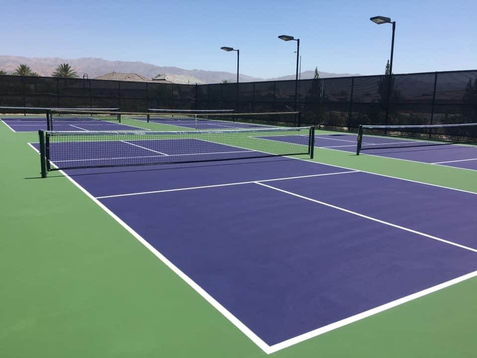  Four pickleball courts are surrounded by a black windscreen that was installed by Palm Springs Tennis Courts. Light poles and mountains are in the background. 