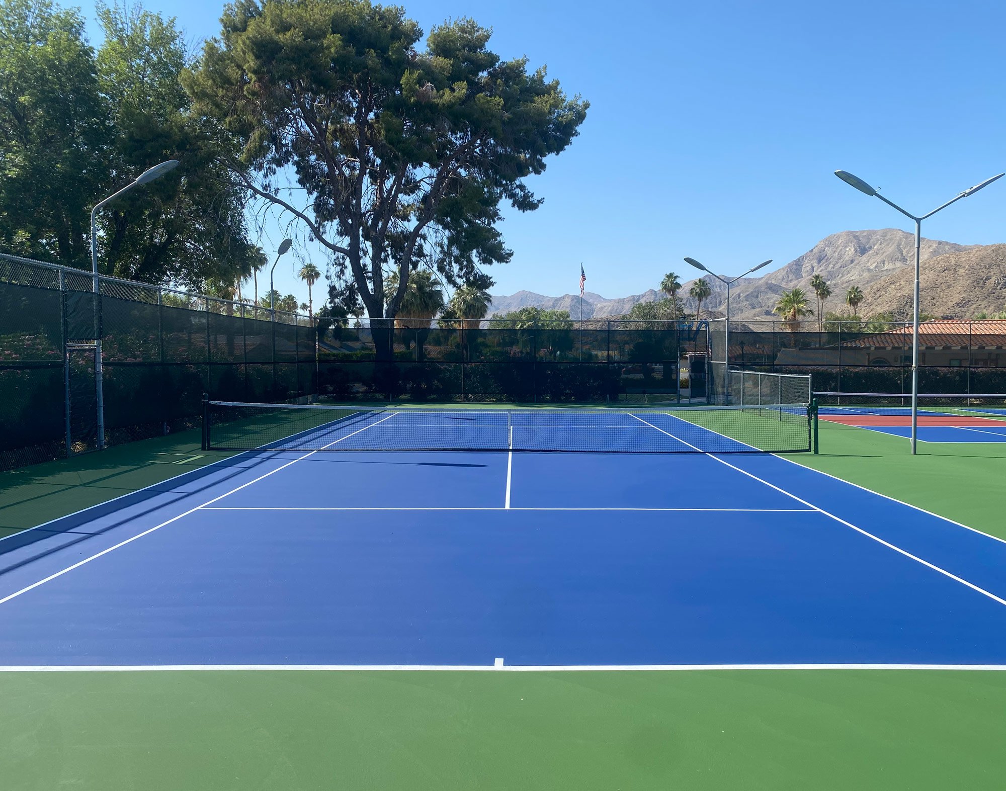  Windscreens surround a multi-court area including tennis court with a blue playing surface surrounded by green surface. It is in a Southern California desert setting. 