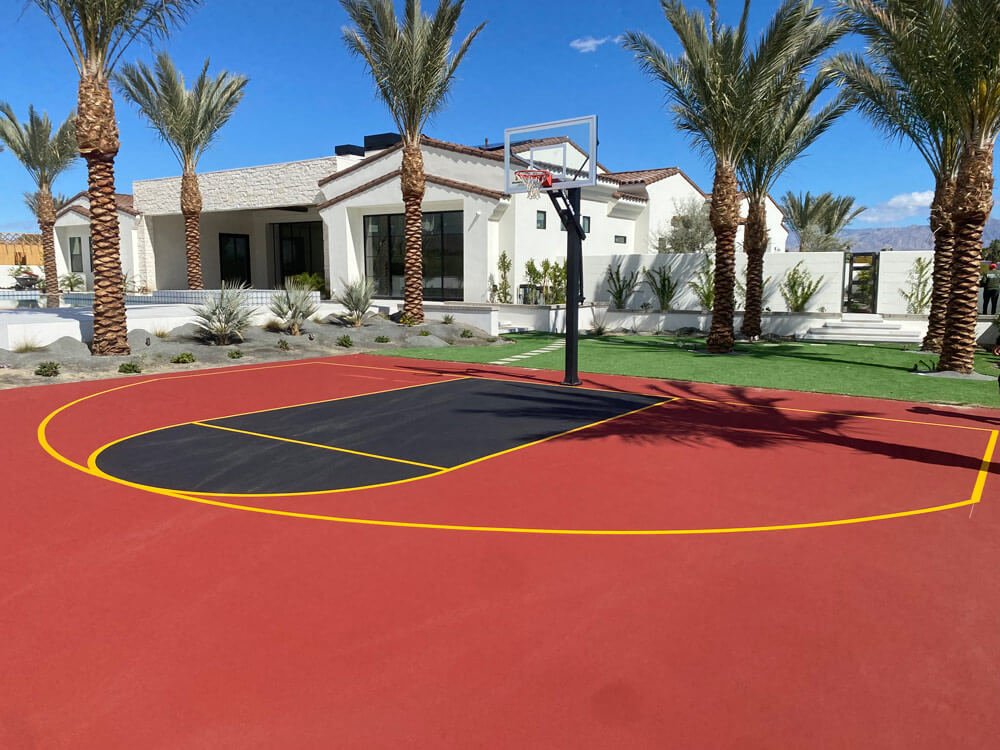  A residential basketball court that has a red surface with a dark blue free throw lane and yellow lines. The court was installed by Palm Springs Tennis Courts. A white house and short date palm trees are behind the court. 