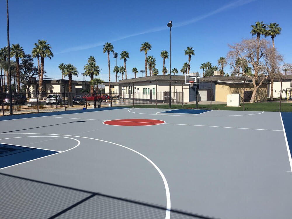  A full basketball court installed by Palm Springs Tennis Courts. The court has a gray surface with a dark blue free throw lane. The center circle is red. 