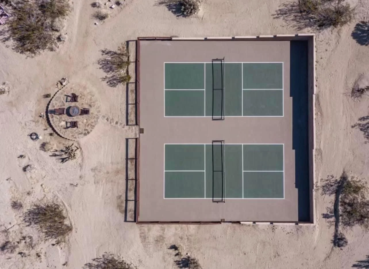 Overhead photo of two pickleball courts on a desert property near Joshua Tree. The courts are green and the surrounding surface is tan. A sitting area with chairs and fire pit is to the left of the courts.