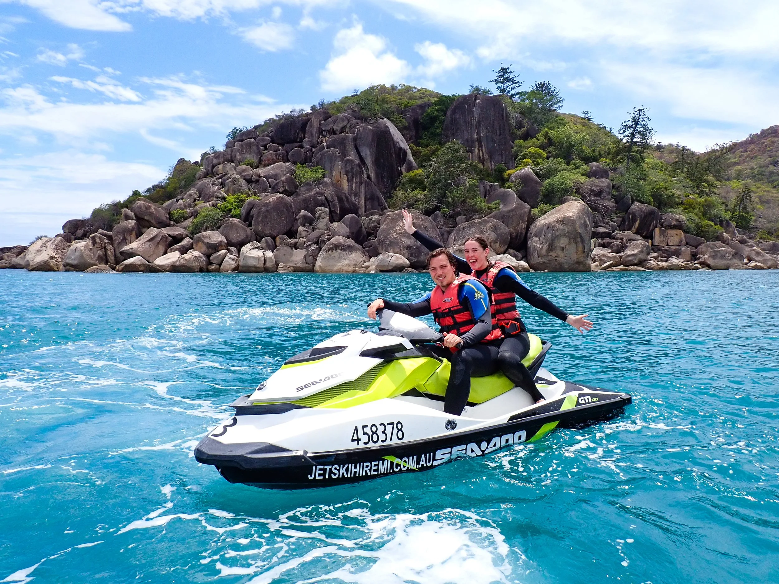 A couple on a jet ski, stopped with arms raised for an excited photo in front of a secluded beach.