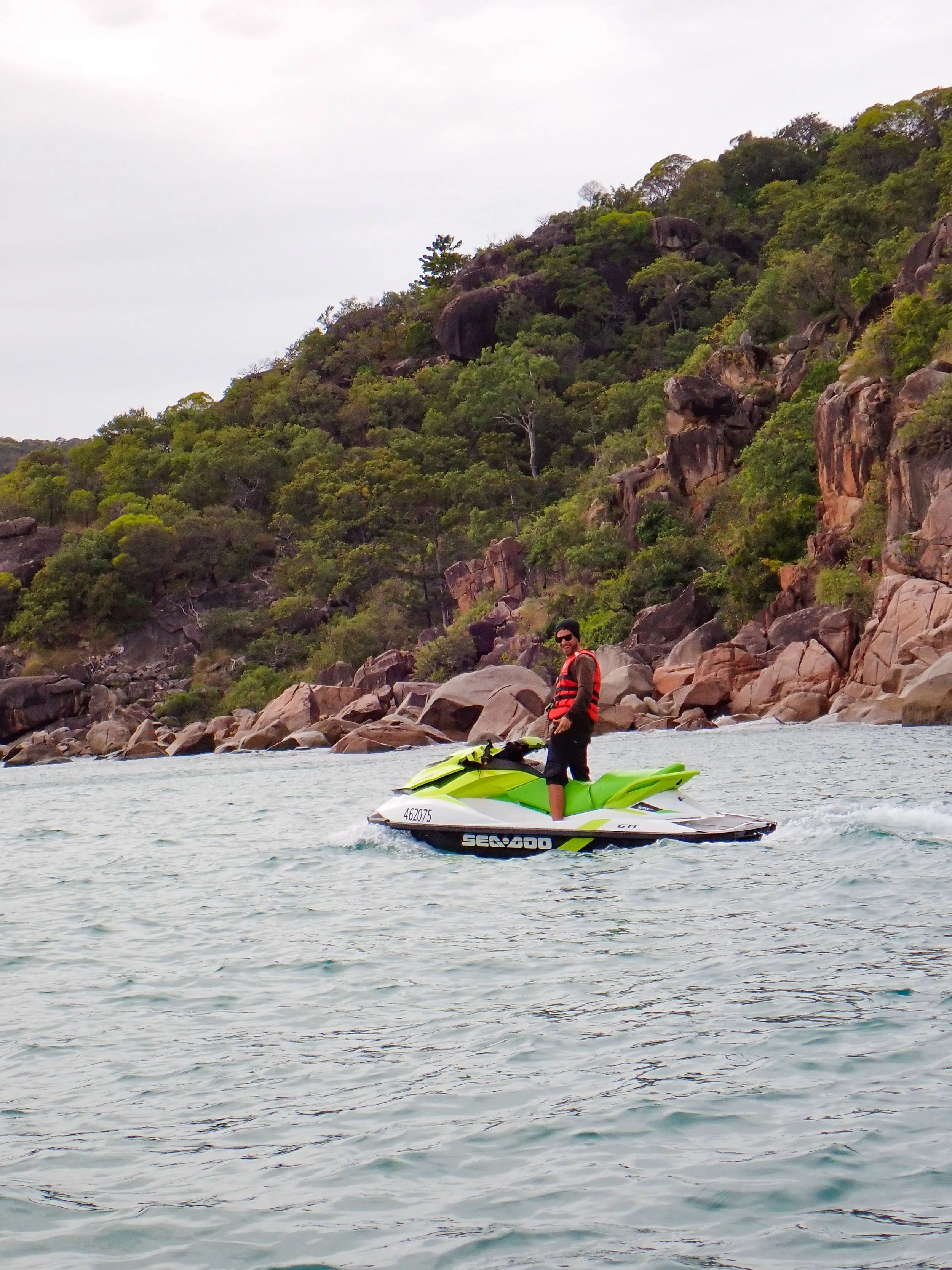 Person in sunglasses and life jacket riding a green and white jet ski on a body of water near rocky shoreline and green trees.