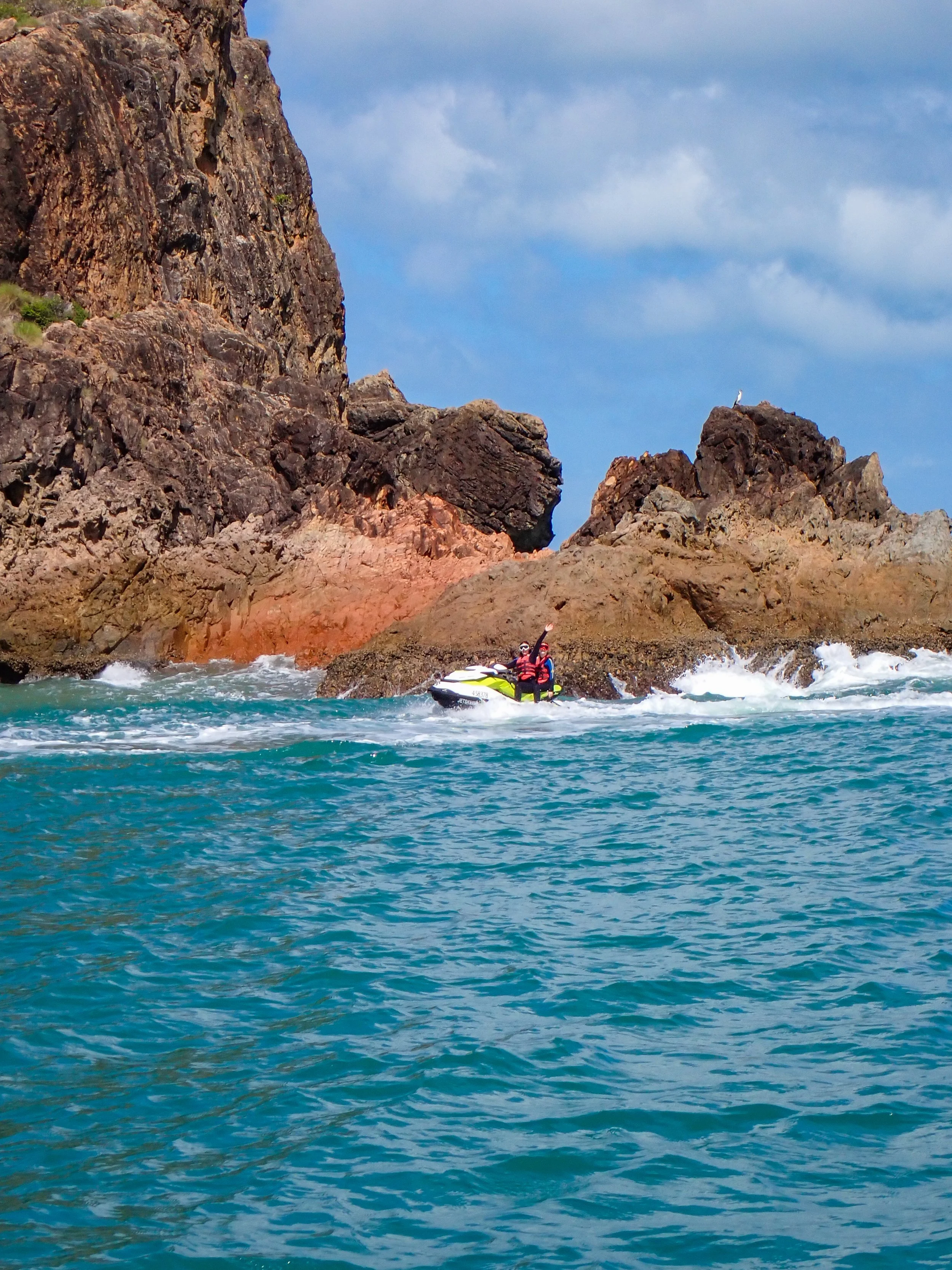 A jet ski with a person waving as they travel along volcanic headlands on Magnetic Island.