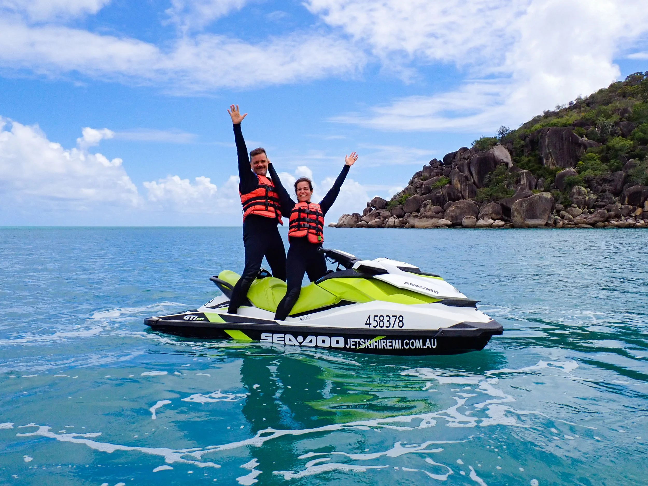 A couple on a jet ski, stopped with arms raised for an excited photo in front of a secluded beach.