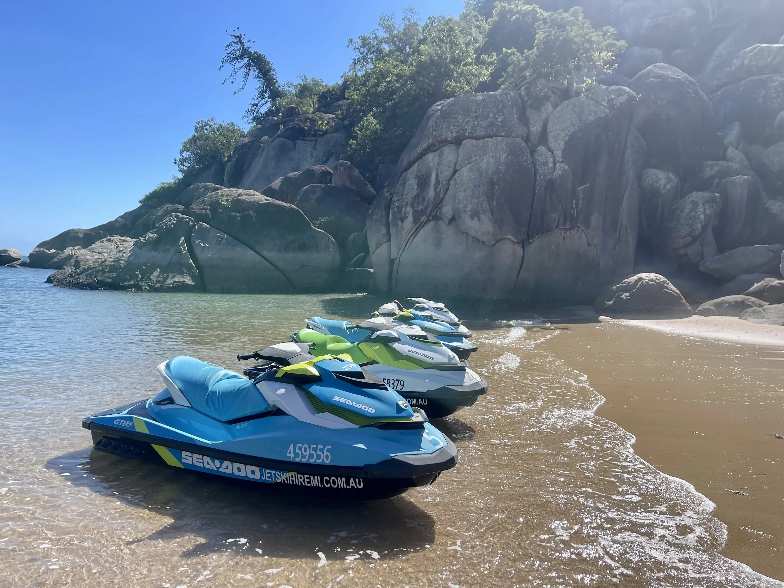 A row of colorful jet skis on a sandy beach near large rocks and greenery, with clear blue skies overhead.