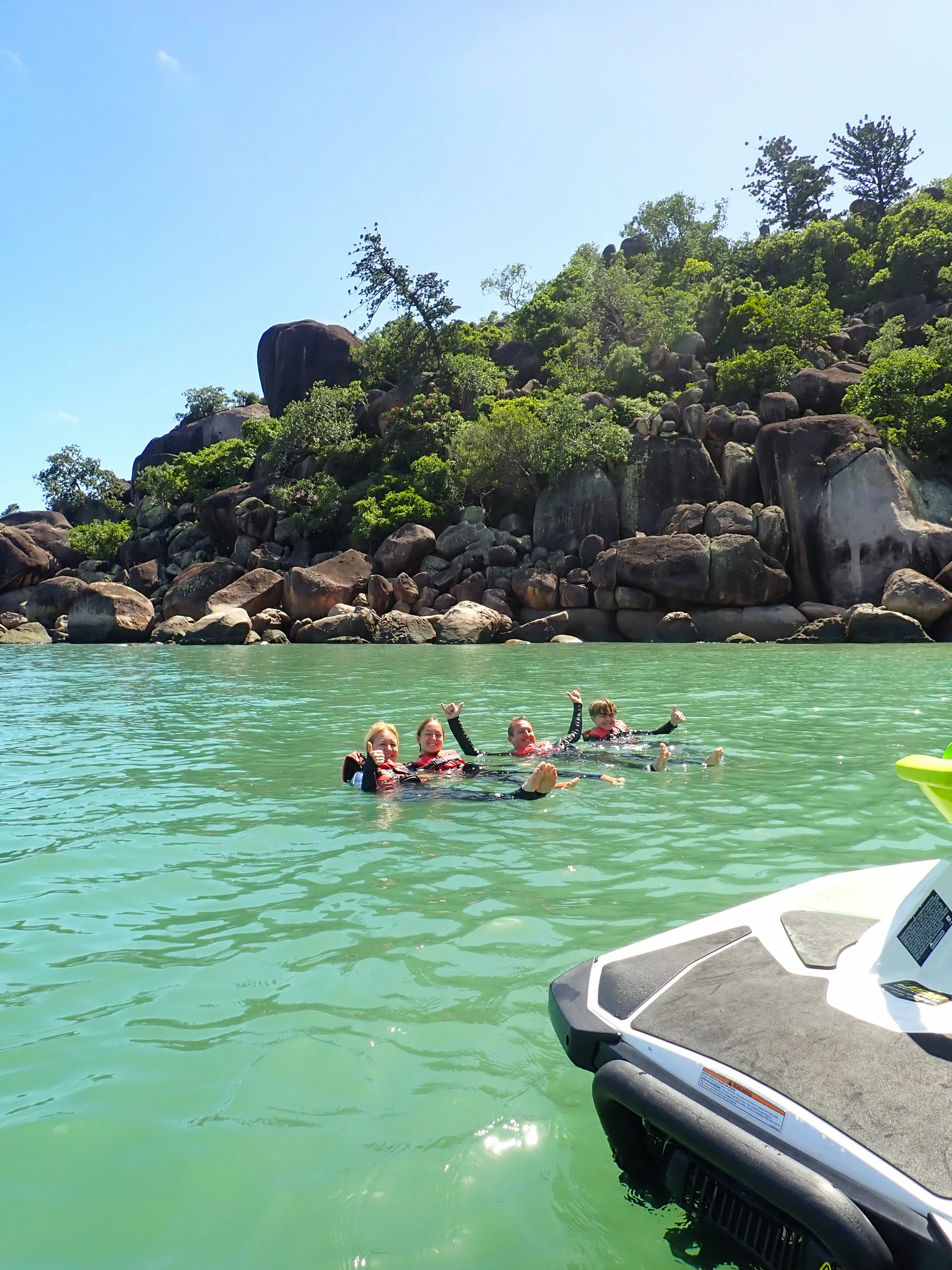A family of 4 swimming in a secluded beach beside their floating jet ski, with a rocky hillside and trees in the background.