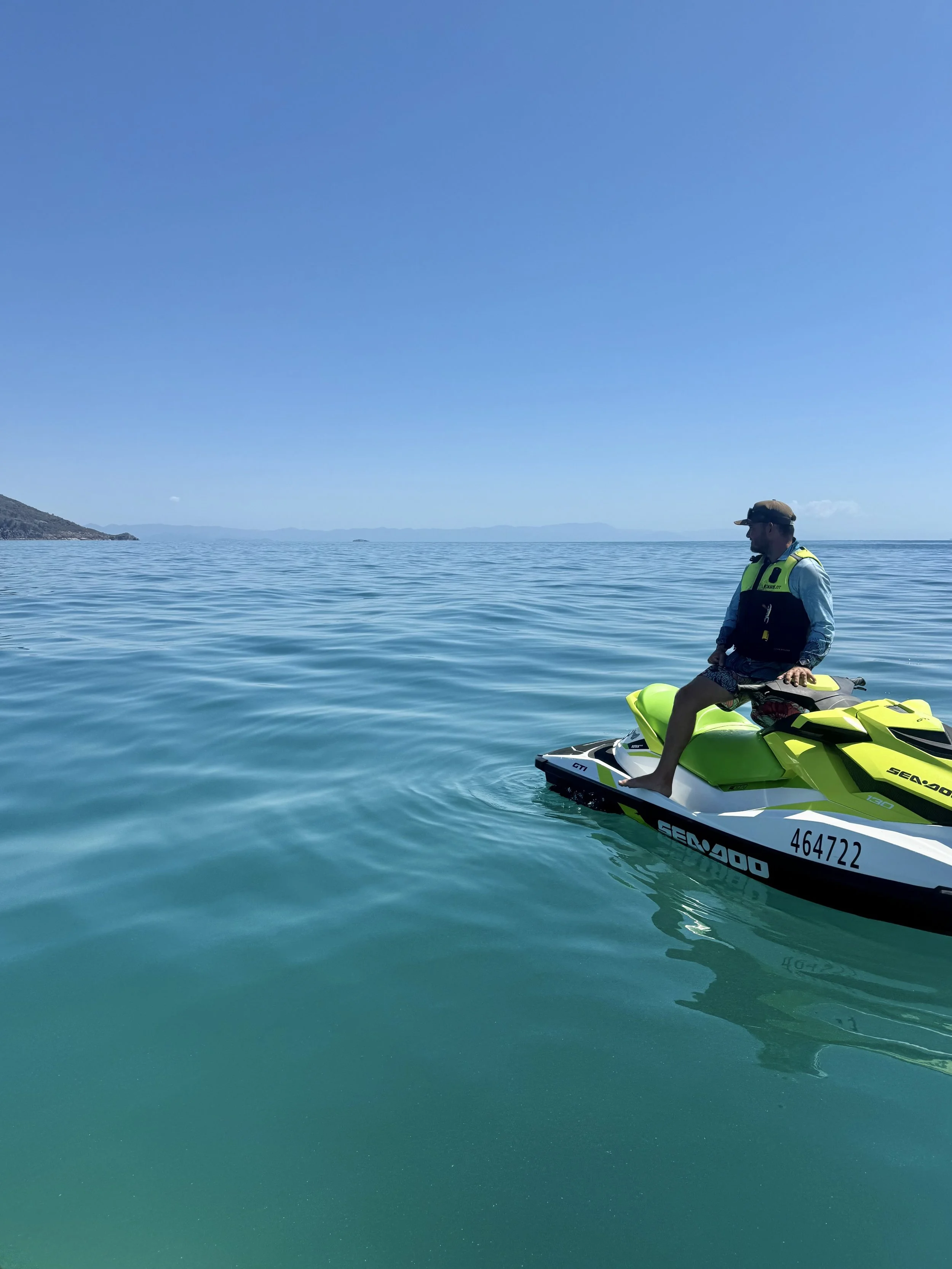 A man riding a green and black Sea-Doo jet ski on calm ocean water under a clear blue sky. He is wearing a life vest, hat, and long-sleeve shirt, with a distant shoreline and mountains in the background.