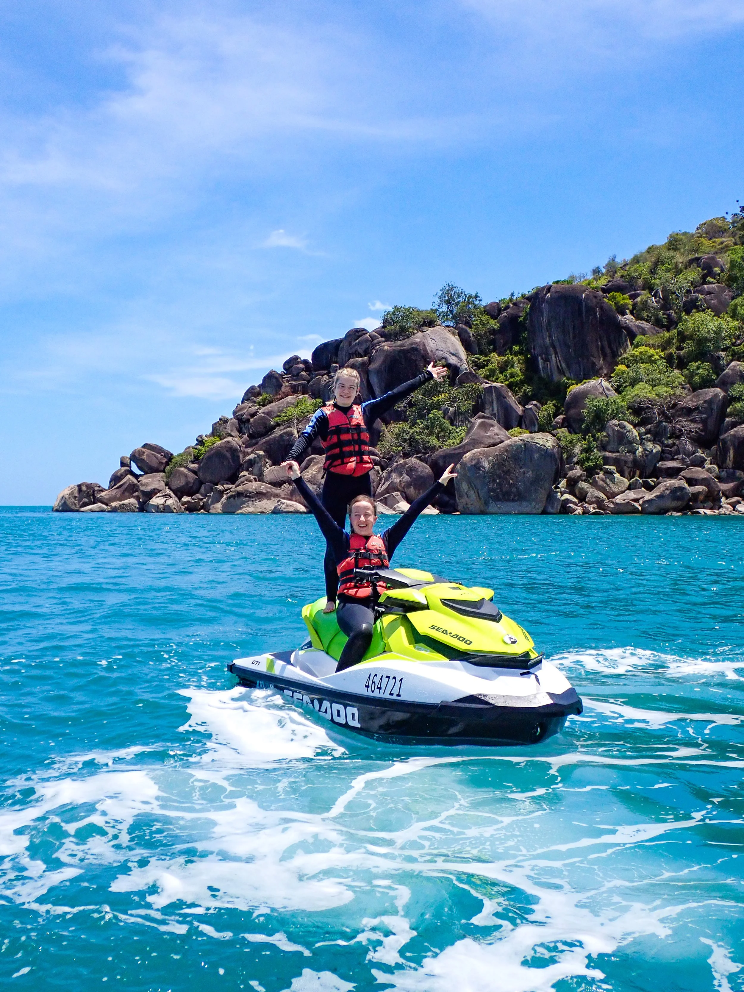 A young pair of friends on a jet ski stopped, standing up with arms raised for an excited photo in front of a secluded beach.