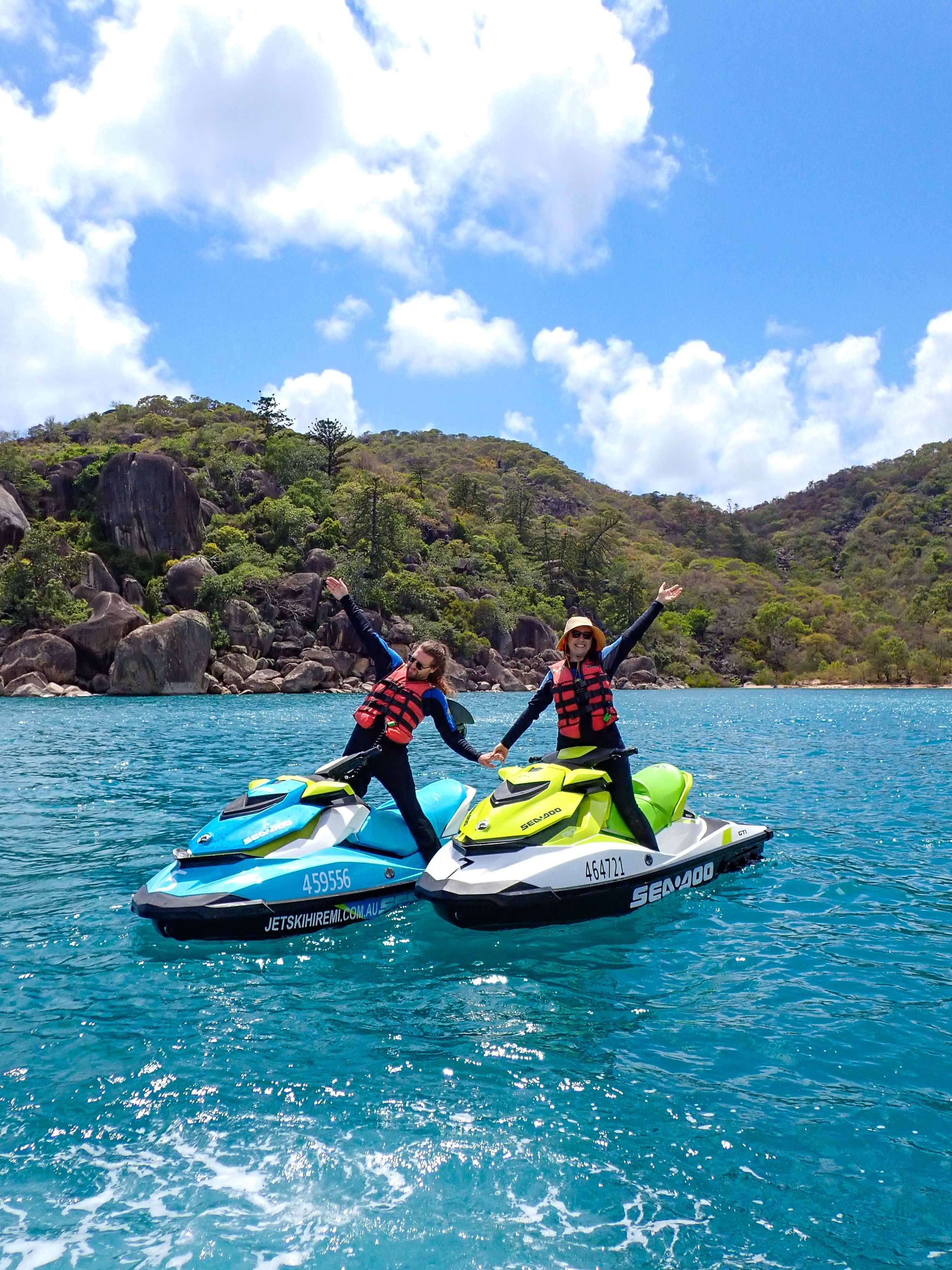 A couple riding a jet ski, stopped with arms raised for an excited photo in front of a secluded beach.