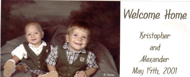 Two young boys sitting on a blanket, smiling, with a welcome sign and names 'Kristopher and Alexander' dated May 19th, 2001.