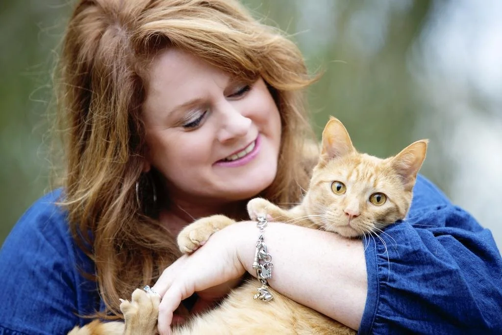 Woman with red hair smiling while holding an orange tabby cat outdoors.
