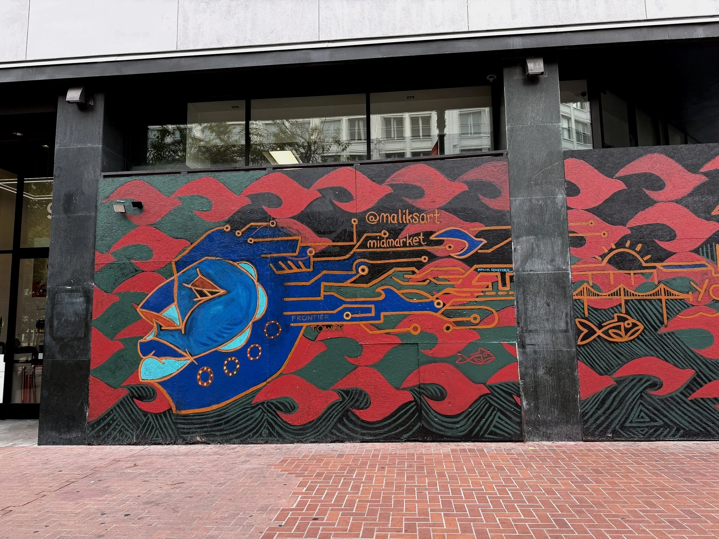 Colorful street art mural on a wall featuring the side profile of a head, stylized fish, abstract waves, and the Instagram username '@maliksart'  in orange. City buildings and trees are reflected in the window above the mural.