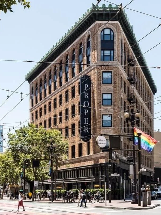 A multi-story brick building with a vertical sign that reads 'THE PROPER' on the corner of Market Street. There are people walking, trees, streetlights, and a rainbow pride flag in front of the building.
