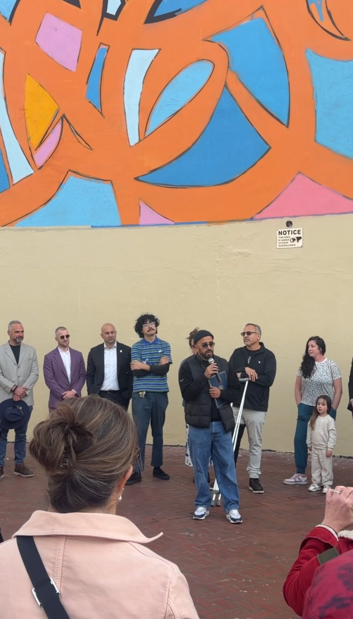 A group of people standing outdoors in front of a beige wall with an orange and blue abstract mural. One person is speaking into a microphone while others listen, including children and adults.