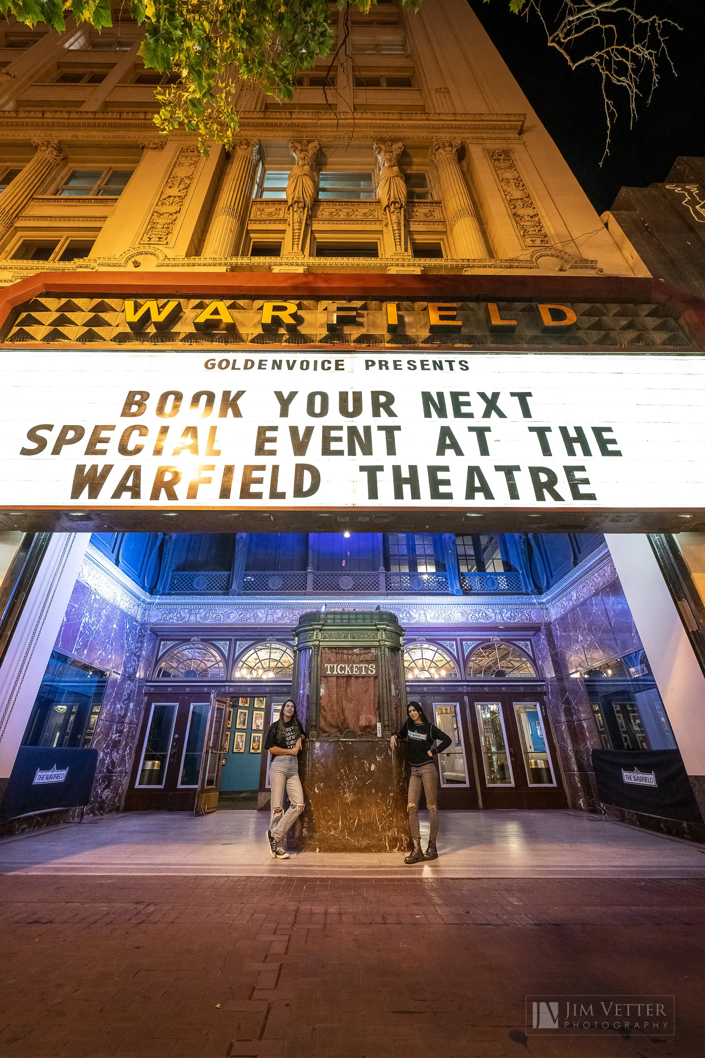 The Warfield Theatre marquee at night advertising a special event booking. The theater's ornate entrance with two women standing in front of a ticket booth is visible beneath the sign.