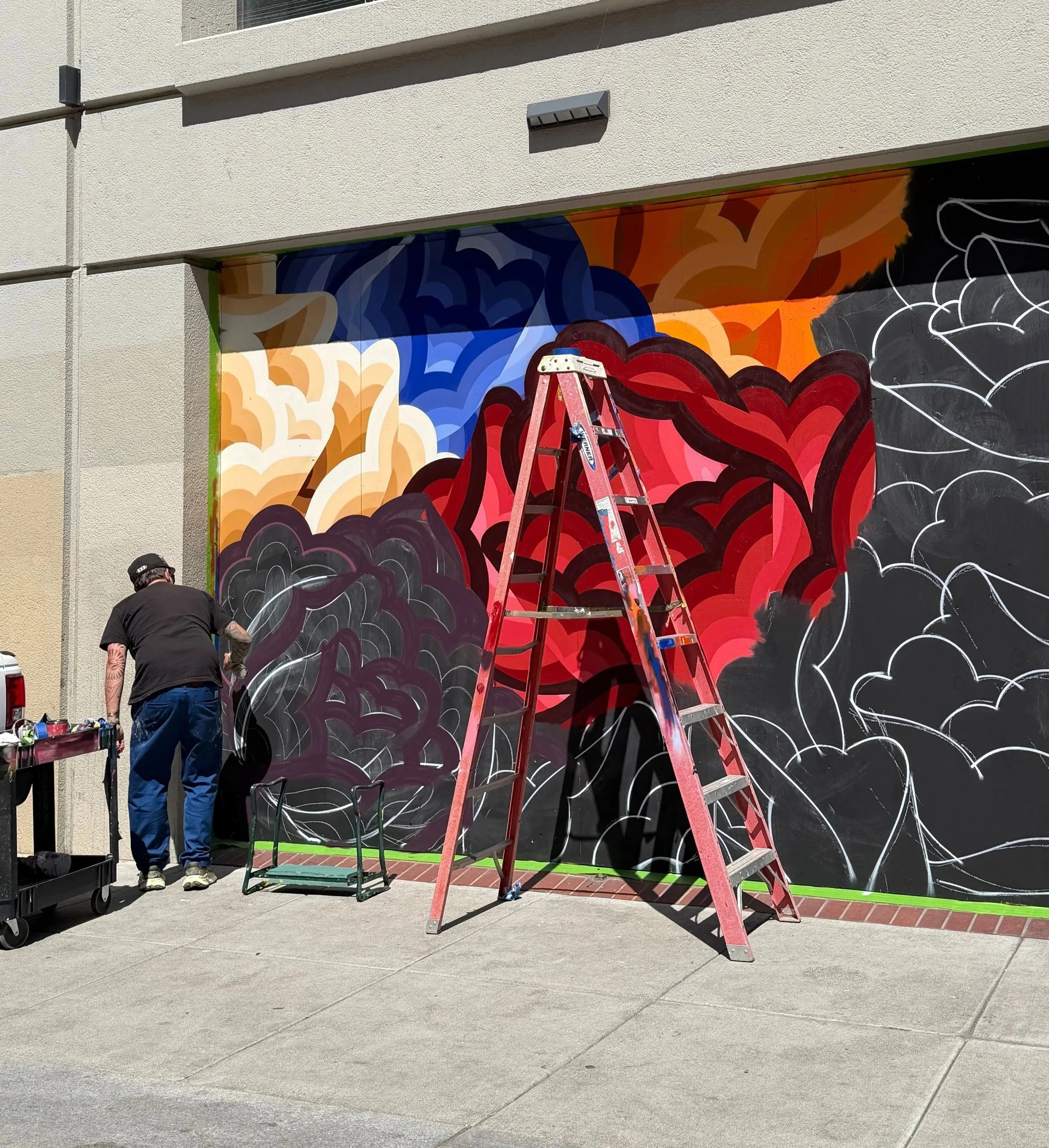 A person with tattoos wearing black clothes is painting a colorful mural on a large black wall outdoors. There are ladders and tools nearby, and the mural features vibrant orange, red, purple, and white roses with gold outlines.