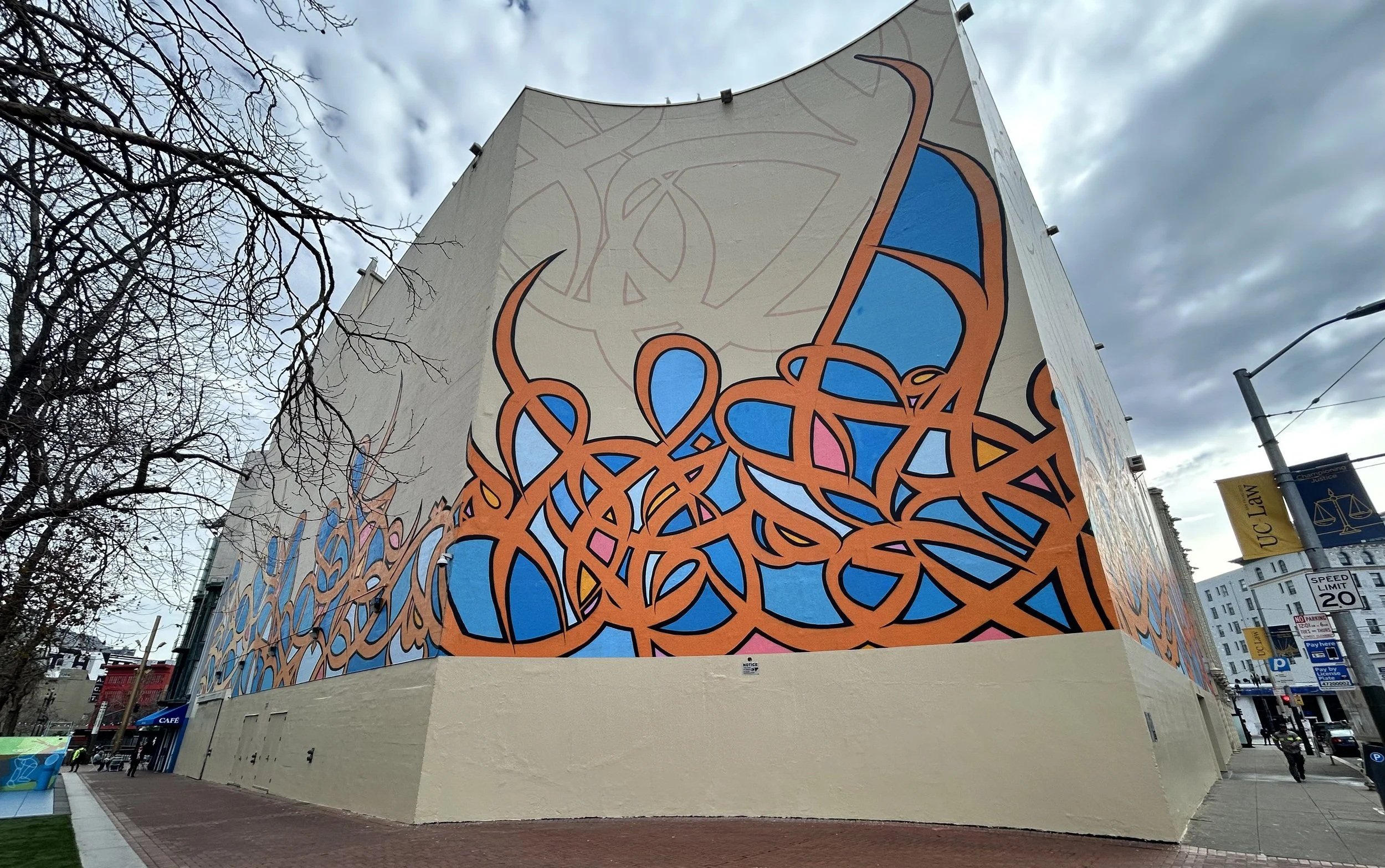 Large building with colorful abstract mural art on the side, featuring intertwining orange lines, blue shapes, and faint outlines of abstract circular patterns. There are leafless trees in the foreground and street signs visible near the sidewalk.
