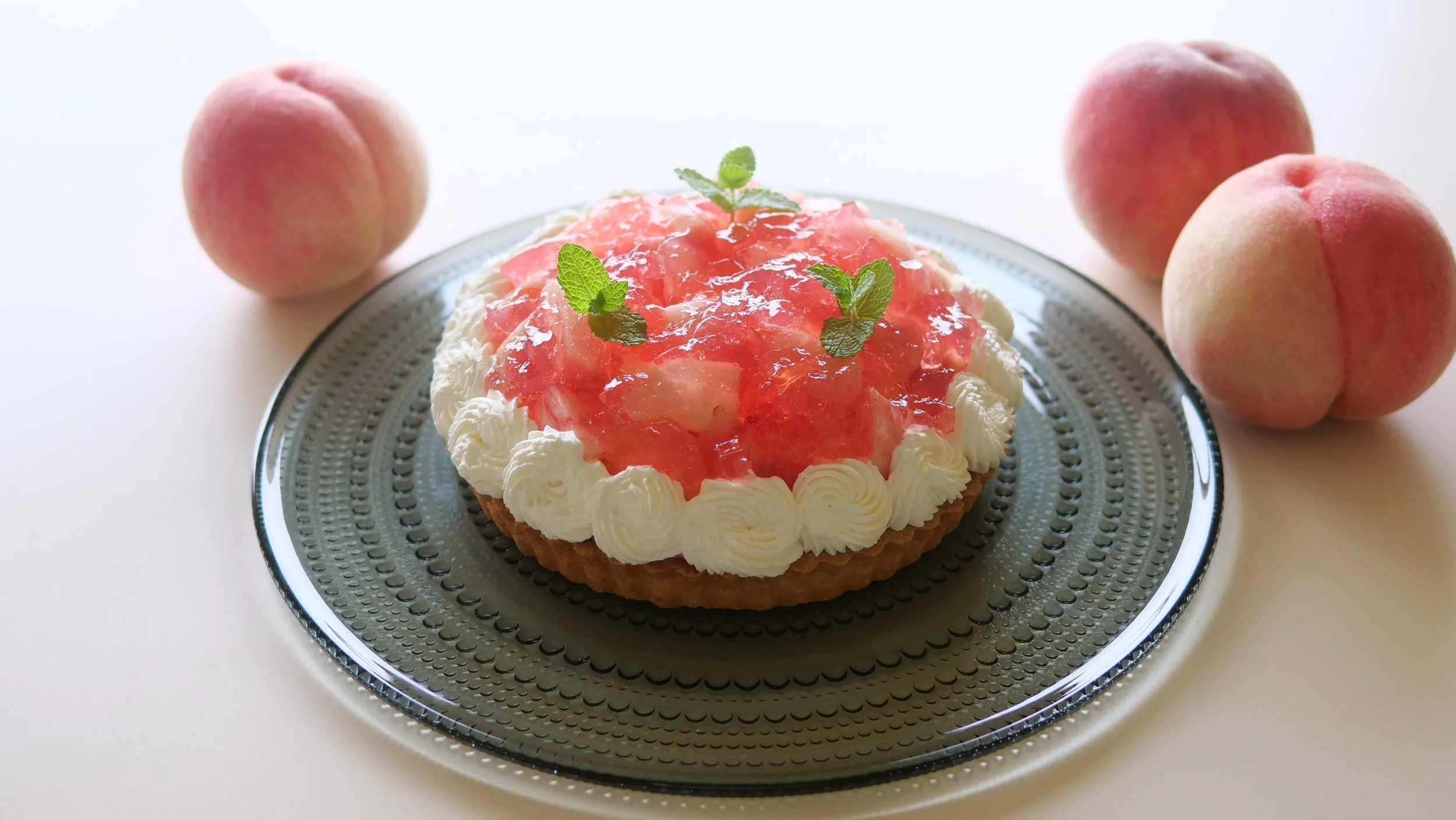 暗めのガラス皿に載った、ホイップクリームとミント、たっぷりのピンク色の桃の果肉で飾られた桃のタルト。