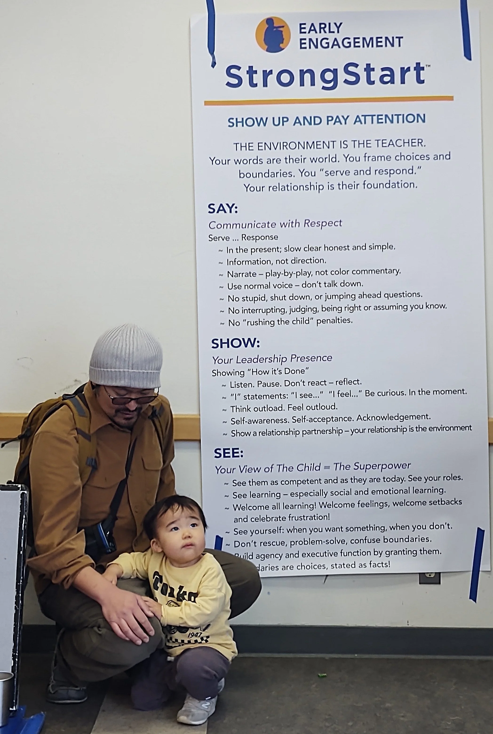 An adult crouching beside a child in front of an educational poster about early engagement and communication strategies titled 'StrongStart.' The poster provides guidance on respectful communication, leadership presence, and viewing children as competent individuals.