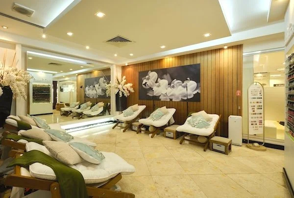 Interior of a spa relaxation area with lounge chairs and cushions, surrounded by decorative plants, soft lighting, and a mirrored wall.