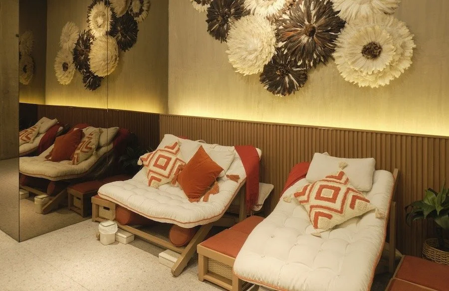 Interior of a spa lounge with reclining chairs, white cushions, and pillows. Warm lighting and wooden panel wall design create a relaxing atmosphere. Orange footstools are placed in front of each chair.