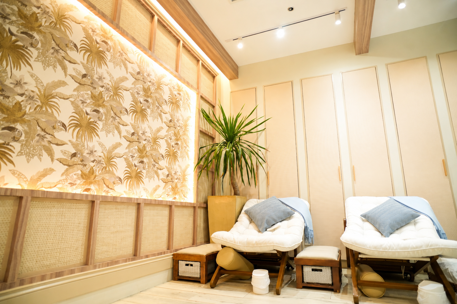 A spa room with four reclining chairs featuring white cushions and green tropical-themed pillows. Each chair has a small wooden footstool. The background includes a stone-textured wall and vertical green plant displays. The ambiance is warm and relax