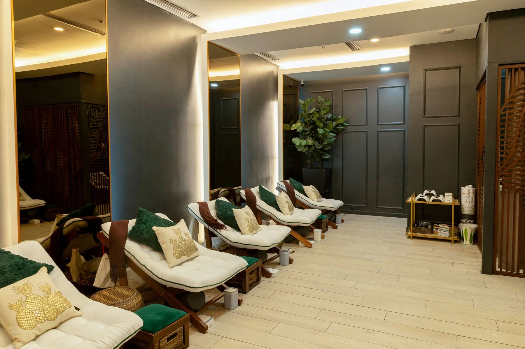 Interior of a modern relaxation lounge with several lounge chairs, cushions with pineapple designs, spa accessories on a small table, and potted plants in a serene, softly lit environment.