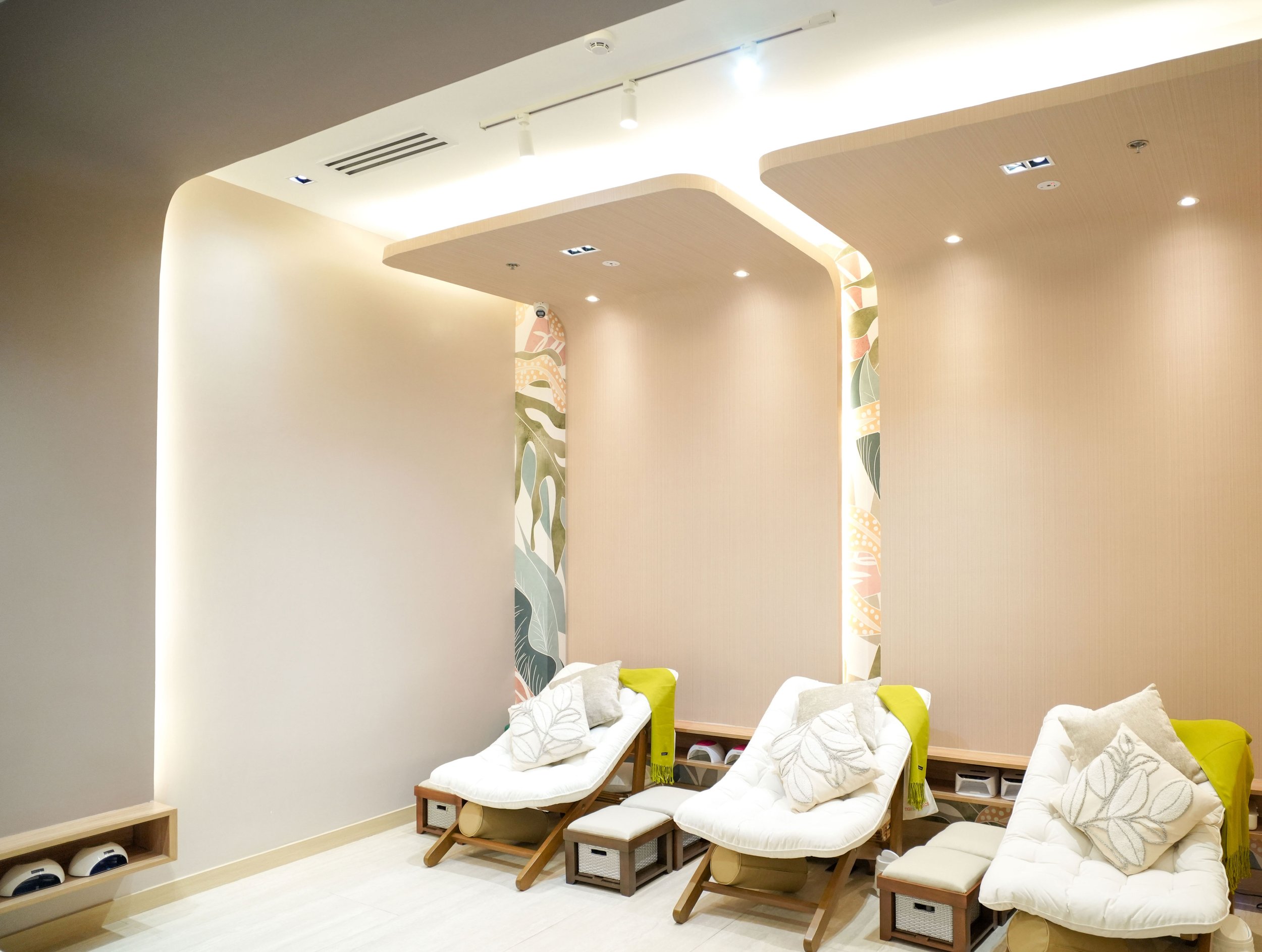 A modern wellness or spa room with several reclined lounge chairs covered with white cushions and pillows, accented with yellow and gray pillows. The room features a curved wooden ceiling with soft white lighting, decorative side panels with abstract leaf patterns, and a minimalist design with light-colored walls and flooring.