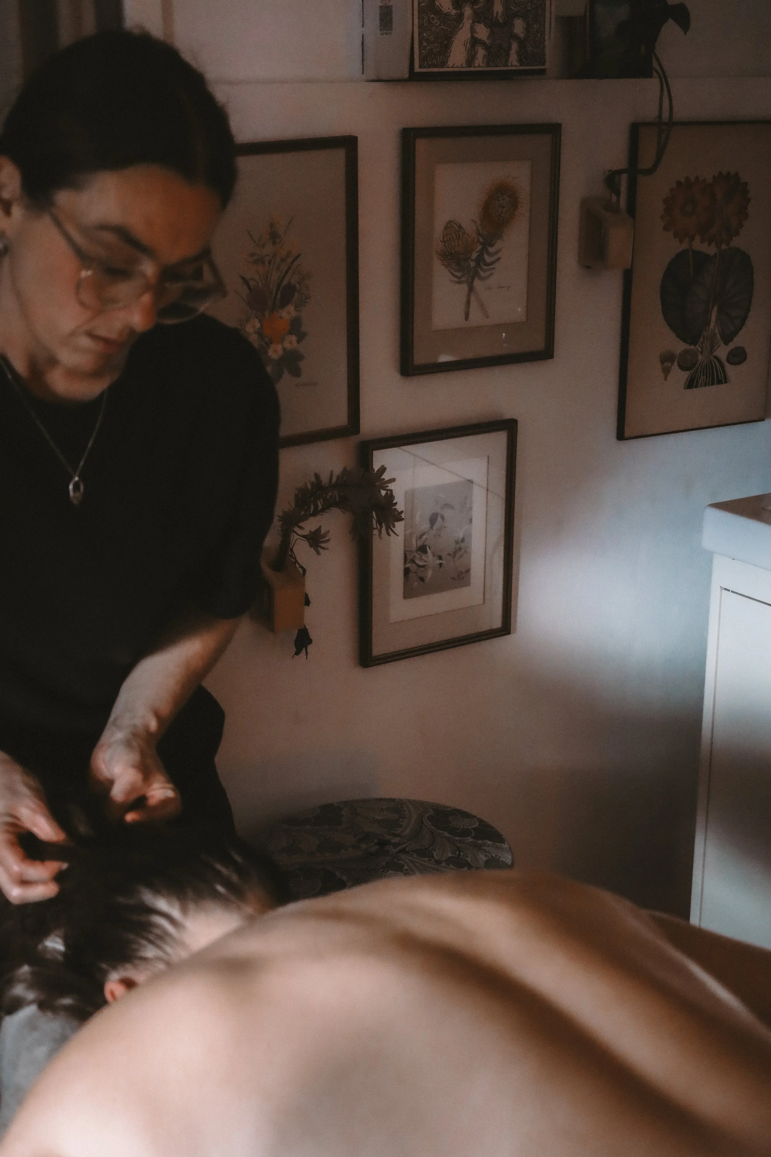 Person lying face down on a massage table while another person gives a massage, with framed botanical artwork on the wall in the background.