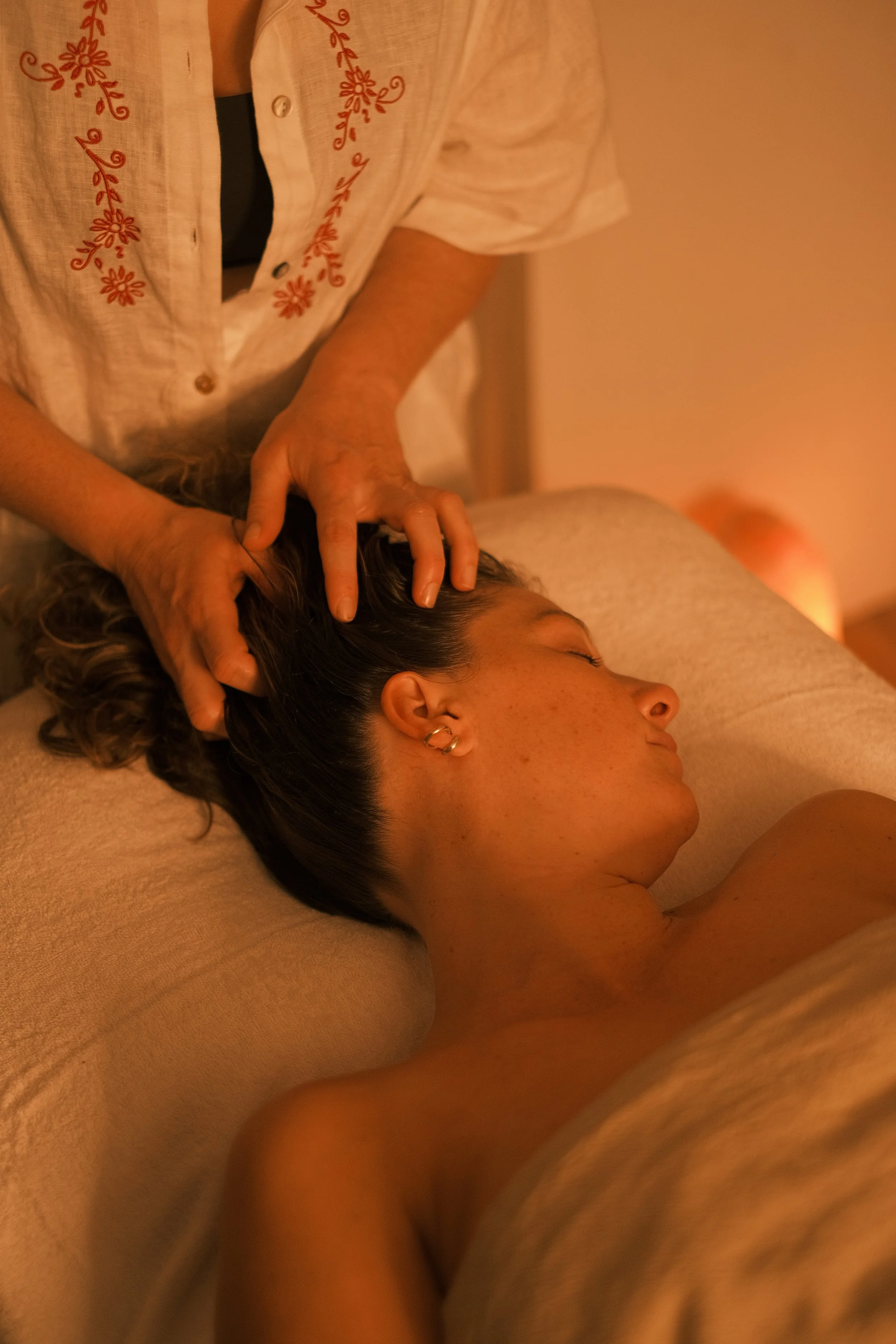 A woman receives a head massage while lying on a massage table in a warm, dimly lit room.