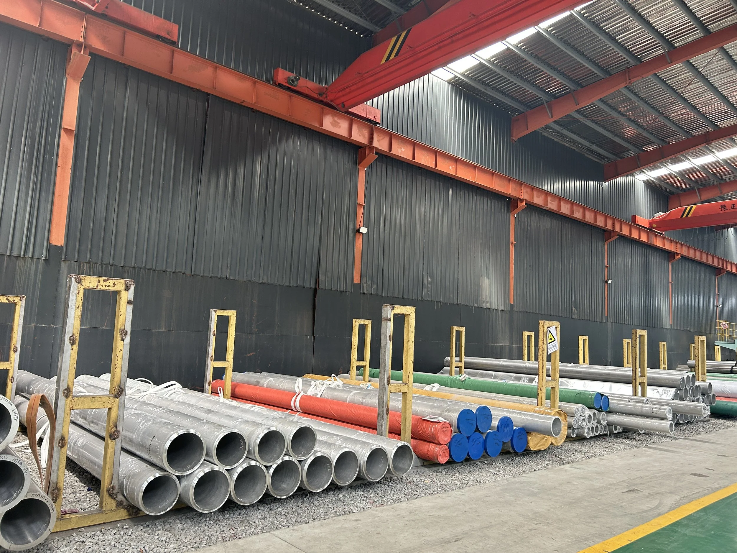 Industrial warehouse with stacked metal pipes in various colors and sizes, including silver, red, green, and blue, alongside yellow safety barriers and a red overhead crane.