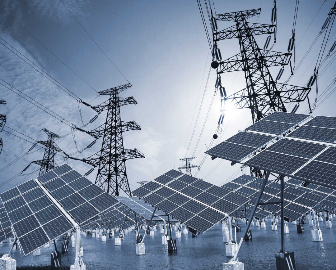 Solar panels and electrical transmission towers on water under a cloudy sky.