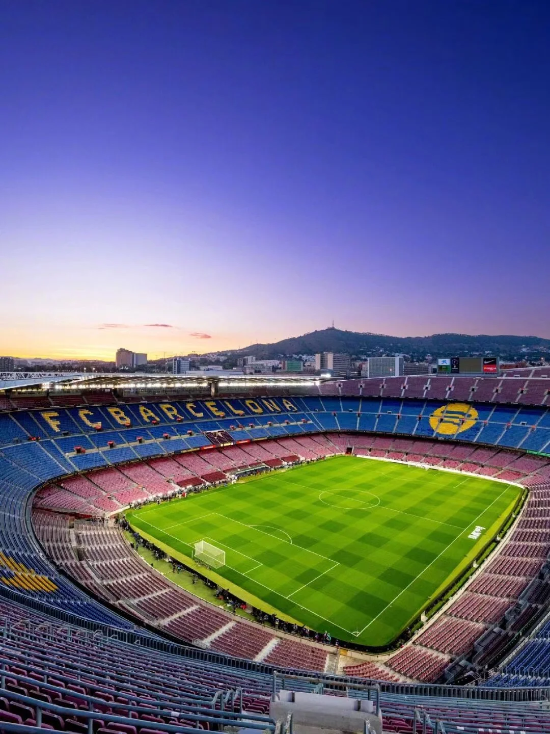 View of Camp Nou stadium in Barcelona at sunset, with the field, seating areas, and cityscape in the background.