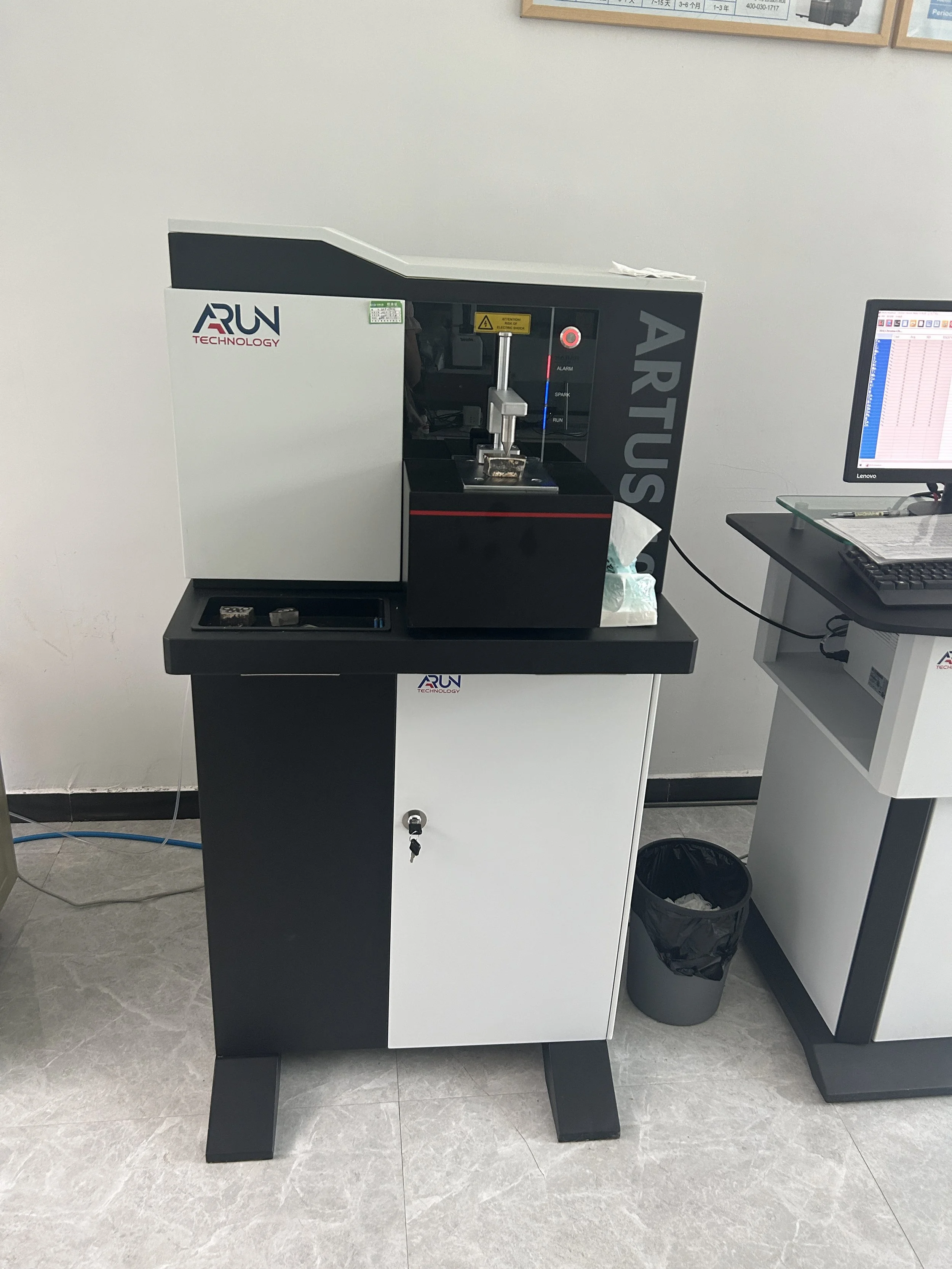 A black and white machine labeled 'ARUN TECHNOLOGY' placed on a black and white cabinet, located in an office or lab environment. To the right, there's a computer monitor, keyboard, and papers on a desk. A small trash can is underneath the desk.