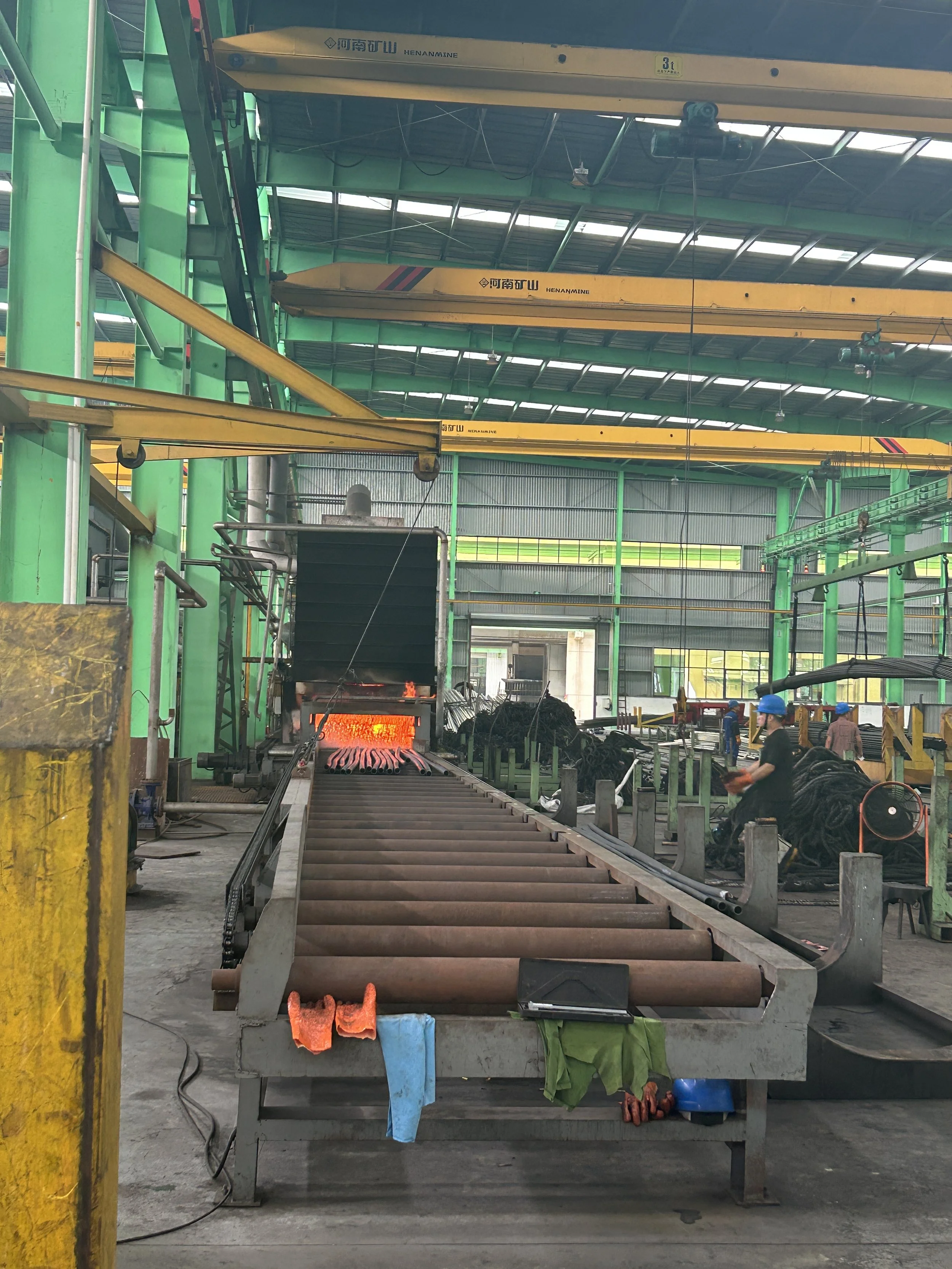 Industrial factory with a furnace burning, surrounded by machinery and workers in blue helmets, amid green and yellow metal structures.