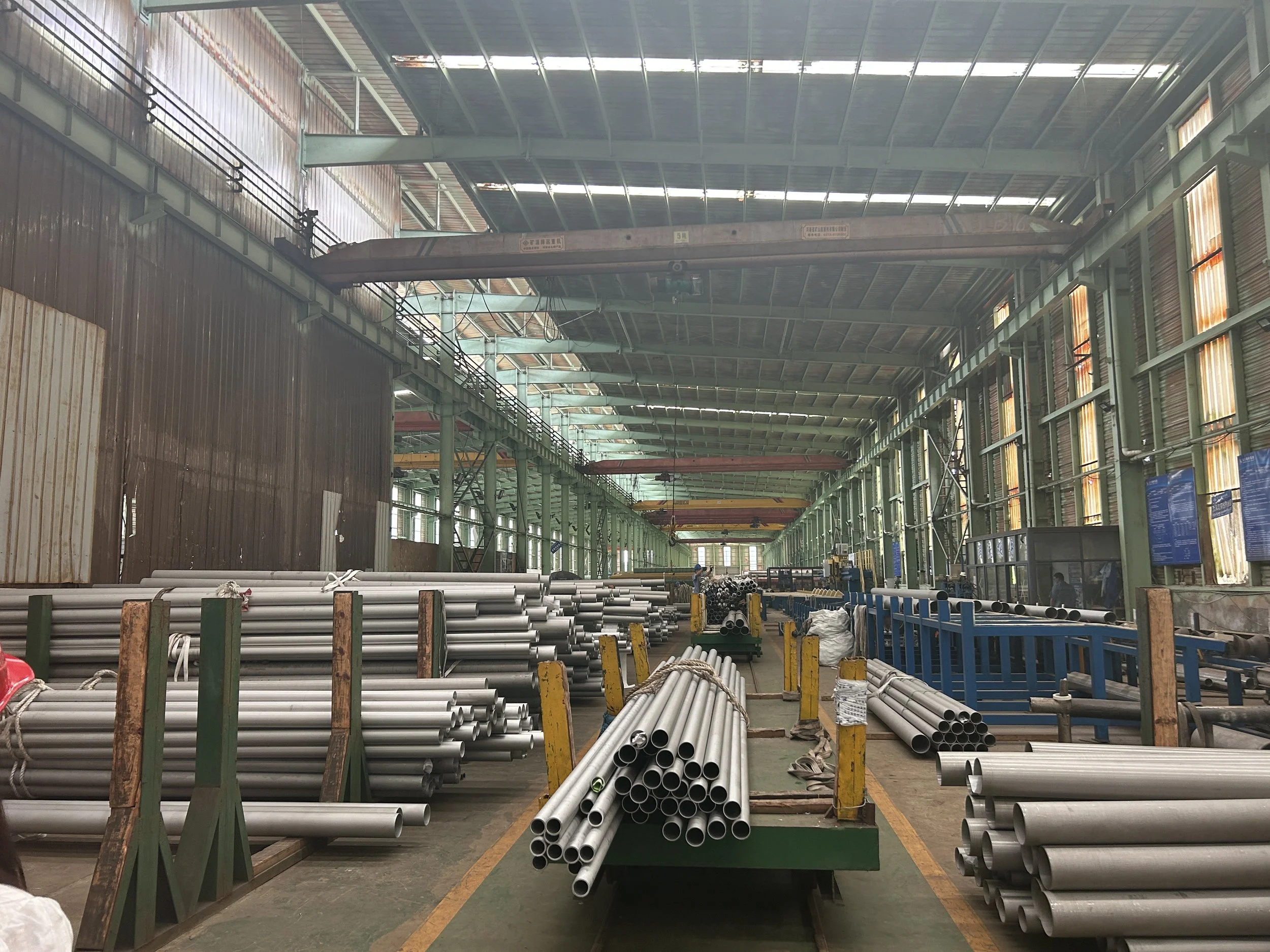A manufacturing warehouse with metal pipes organized on green racks and tables, overhead cranes, and large windows letting in natural light.
