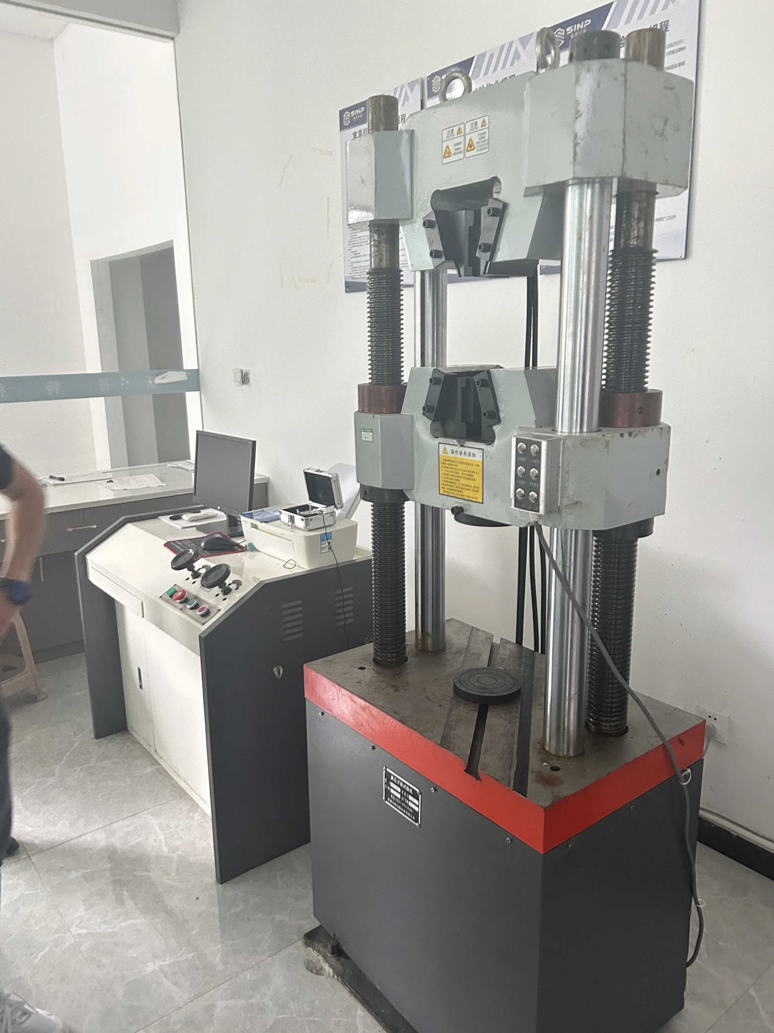 A large industrial compression testing machine with a control panel on a nearby table.