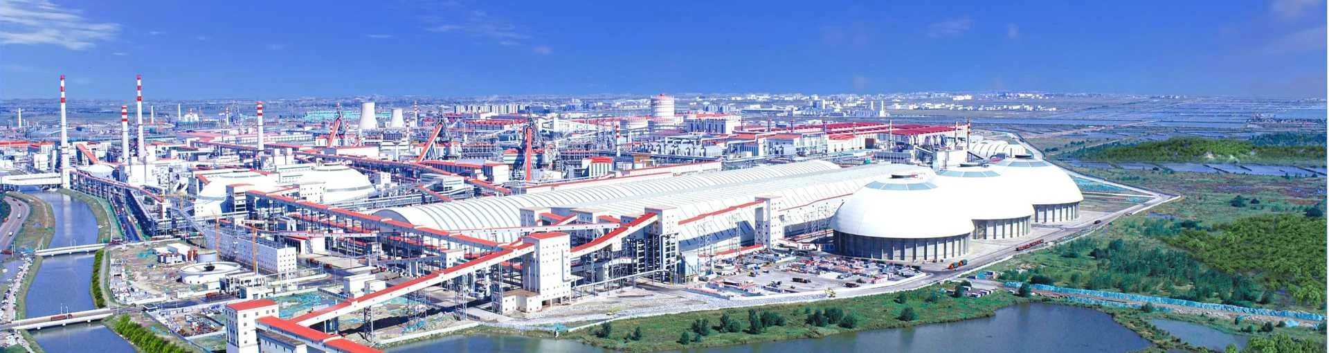 A large industrial complex or power plant with multiple buildings, smokestacks, and cooling towers, situated near water bodies under a clear blue sky.
