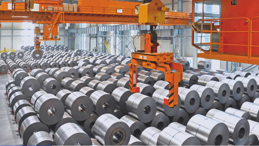 Industrial factory with rows of large metal coils and a robotic arm hanging from an orange overhead crane.