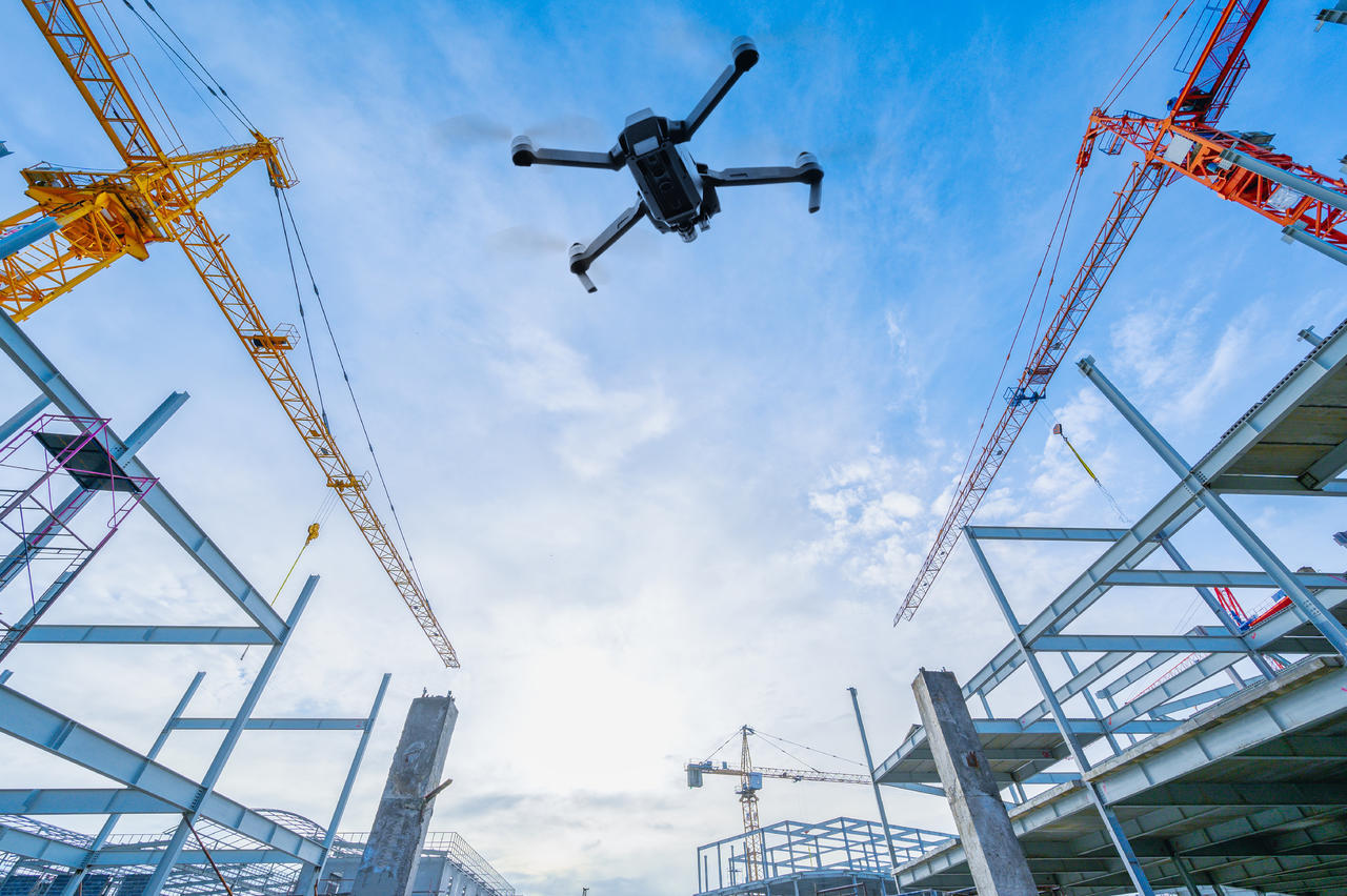 Construction site with steel framework, two cranes, and a drone flying overhead against a partly cloudy sky.