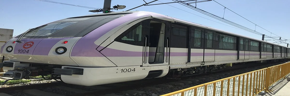 A passenger train on railway tracks with a purple and white exterior, overhead electric wires, and a yellow safety railing in the foreground.