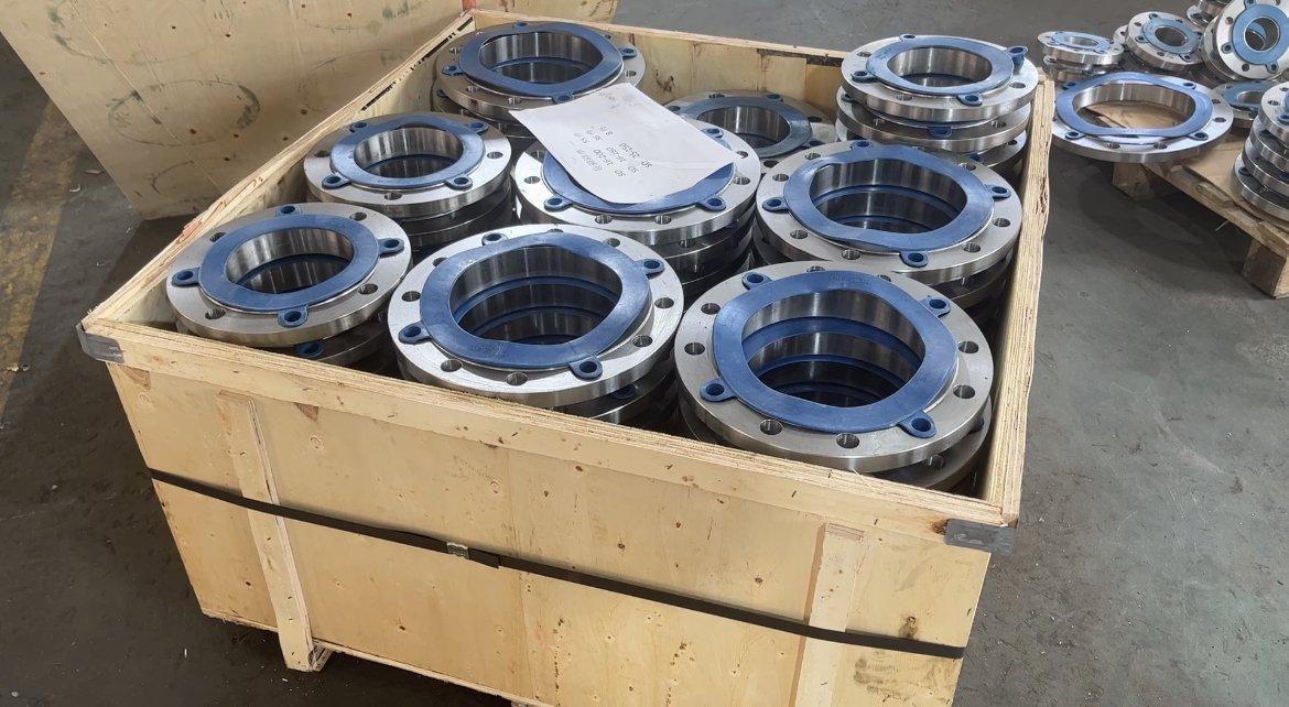 A large wooden crate filled with ASTM Stainless flanged pipe fittings stacked in a workshop or manufacturing area.