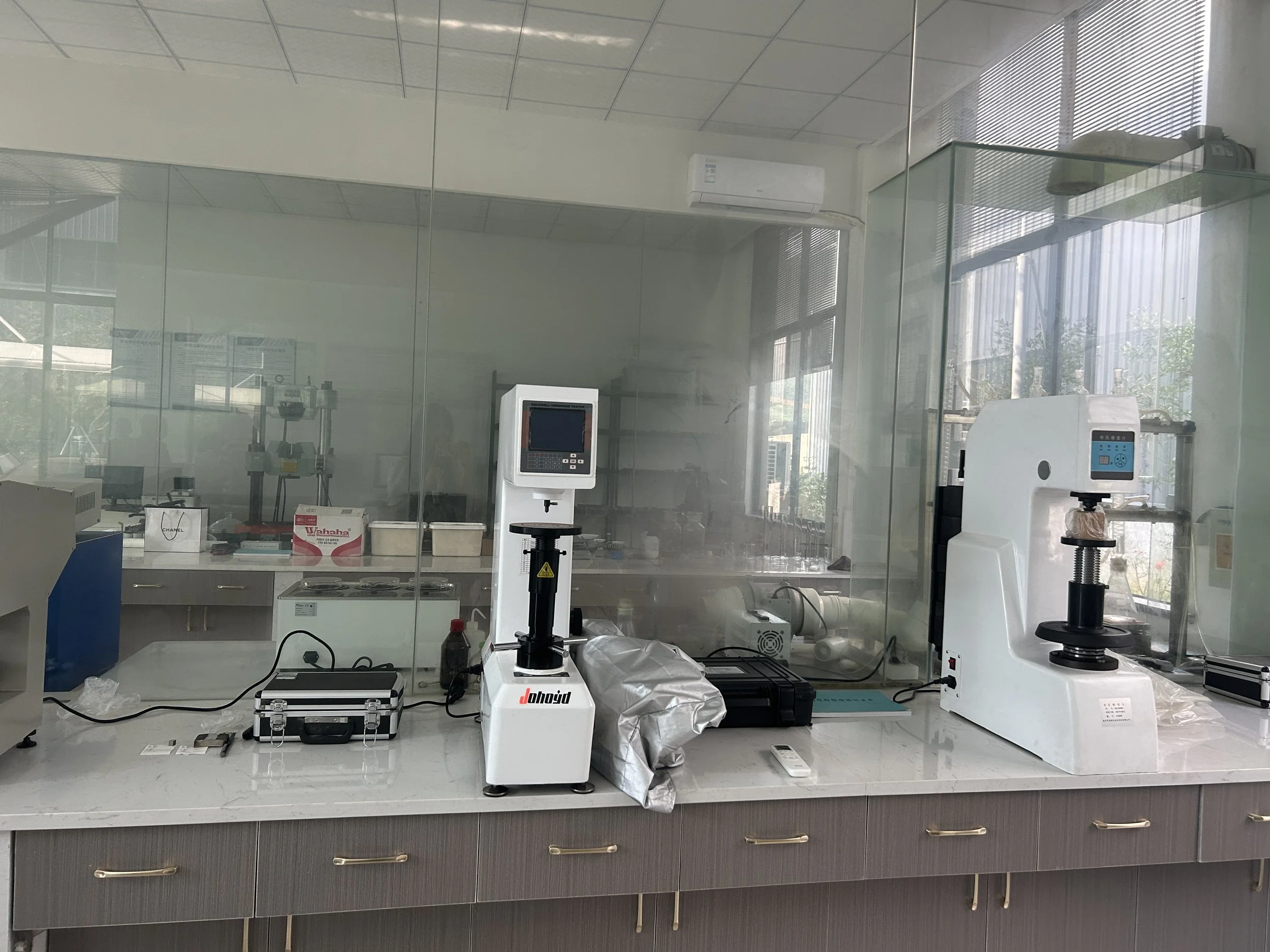 Laboratory workspace with scientific equipment behind glass, including machines and a workbench, with sunlight coming through large windows.
